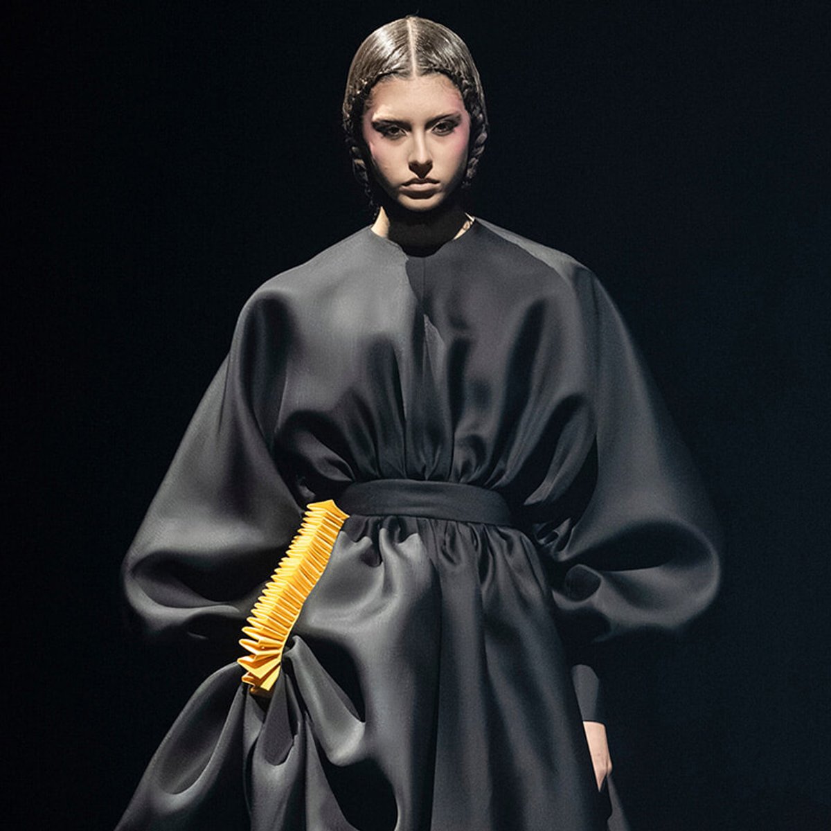 Model wearing a dark, oversized gown with dramatic sleeves and a bright yellow comb-like accessory at the hip.