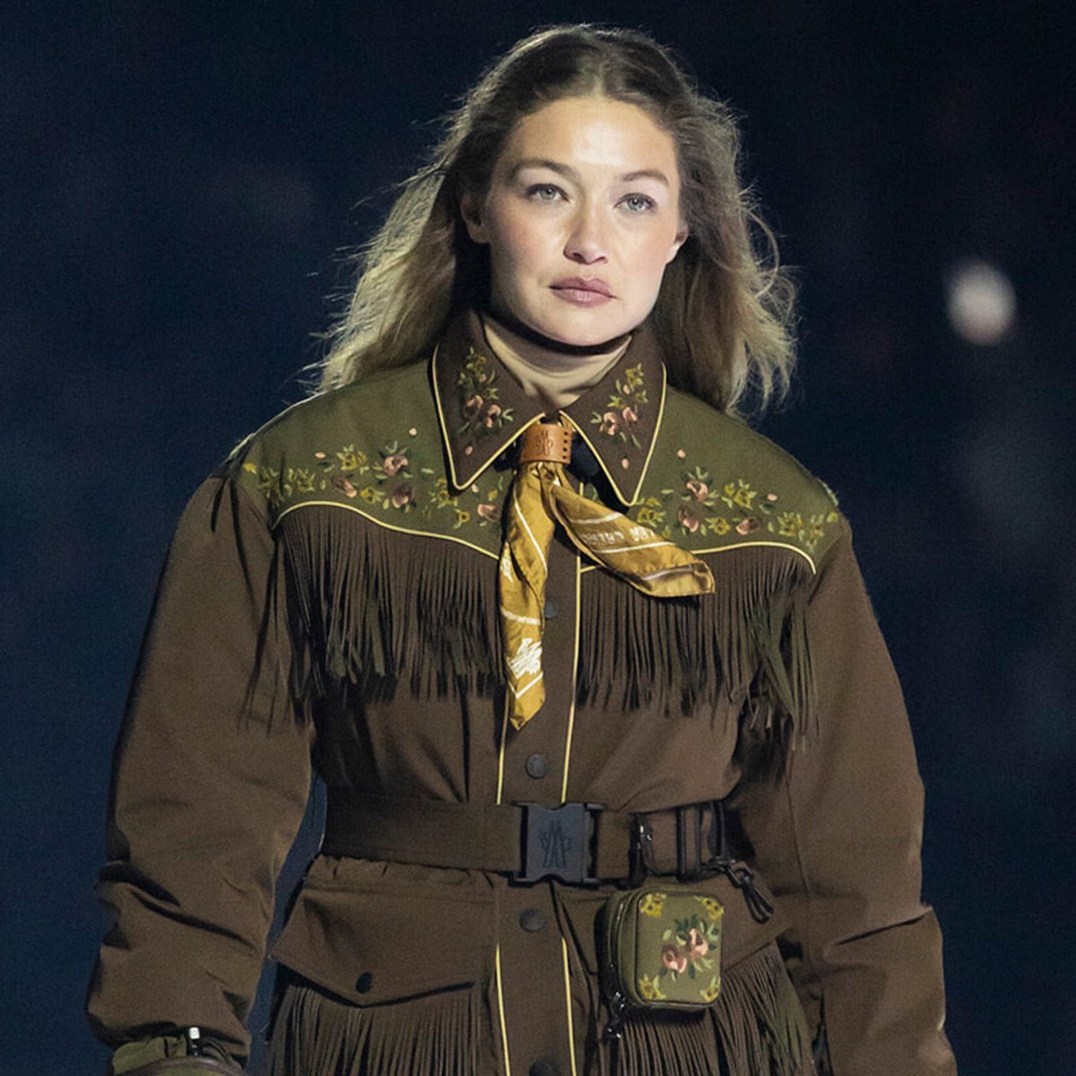 Model walking a runway in a brown fringe Western-style jacket with floral embroidery, yellow scarf, belt, and small handbag.
