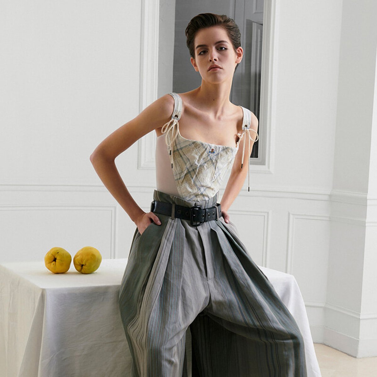 Fashion model posing in a white room, wearing a plaid bustier top with wide shoulder straps and oversized pleated gray trousers held by a black belt; lemons on a table beside her.