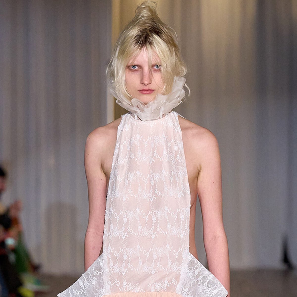 Model walking the runway in a sheer white lace halter dress with a high ruffled neck collar.
