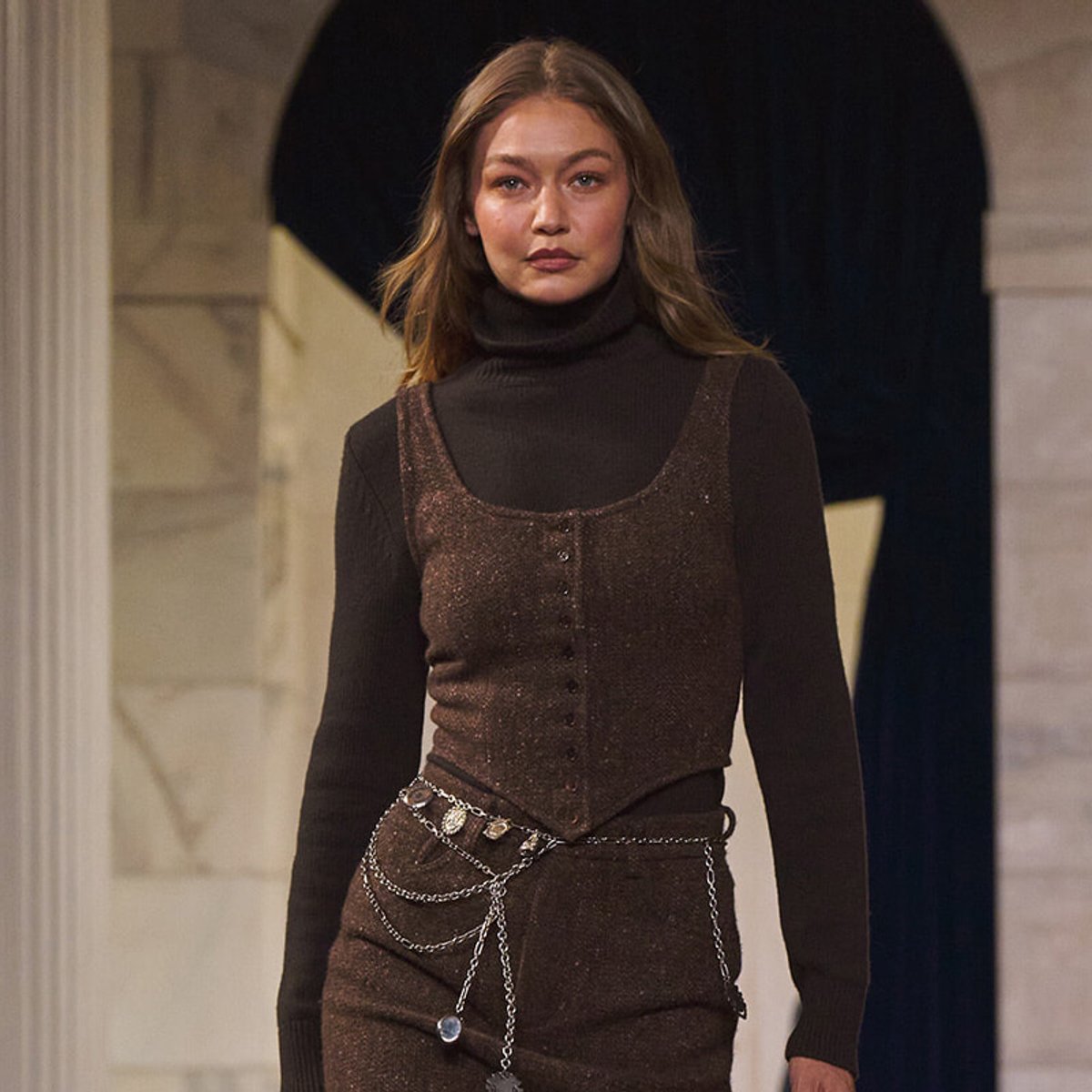 Young woman in a dark turtleneck and a brown glittery sleeveless top with a layered silver chain belt.