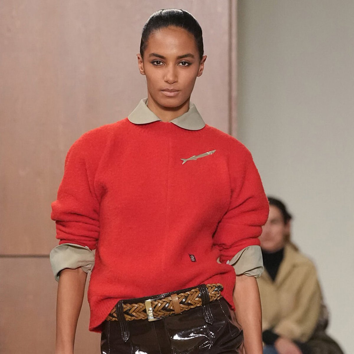 Female model on a runway wearing a red knit sweater with a light gray collar, rolled-up sleeves, and a woven belt with dark pants.
