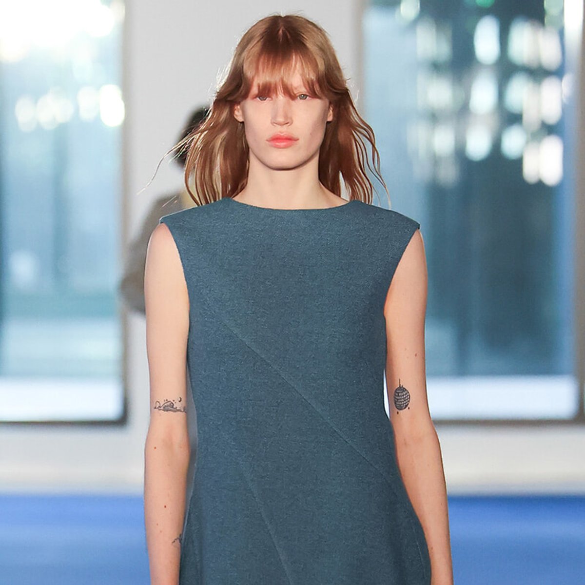 Model walking a runway in a teal sleeveless dress with blunt red hair and small forearm tattoos.