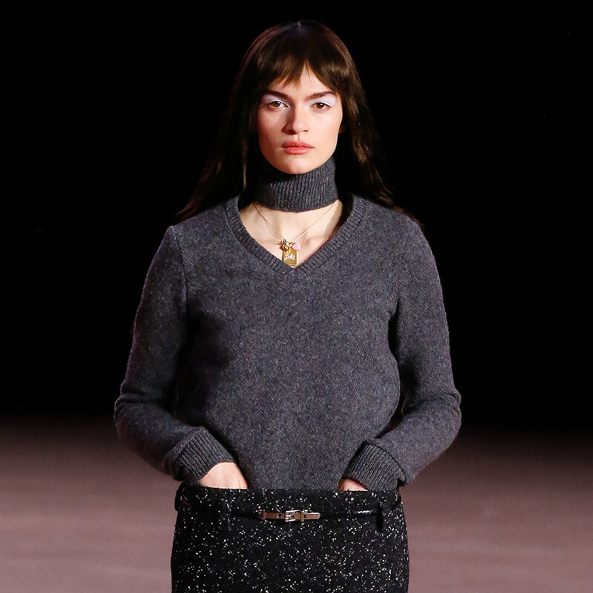 Woman model wearing a dark gray knit turtleneck with hands in pockets on a runway, girl-next-door styling and a gold pendant necklace visible