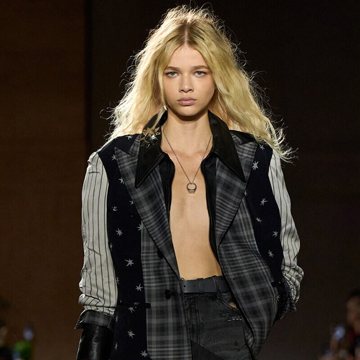Model walks the runway wearing a patchwork plaid jacket with star details, open at the chest and a black belt visible at the waist.