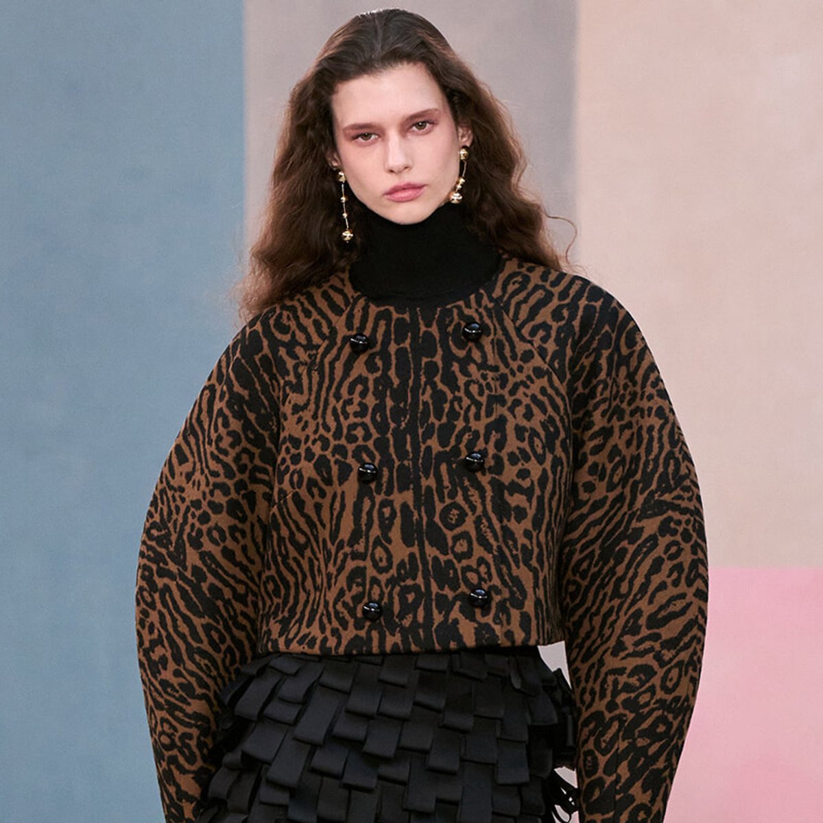 Model wearing a brown leopard-print coat over a black turtleneck, with gold statement earrings, posing against a pastel backdrop.