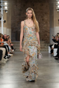 Model on a runway wearing a long, glittering floral sequin gown with cascading ruffles and a pale pink quilted purse.