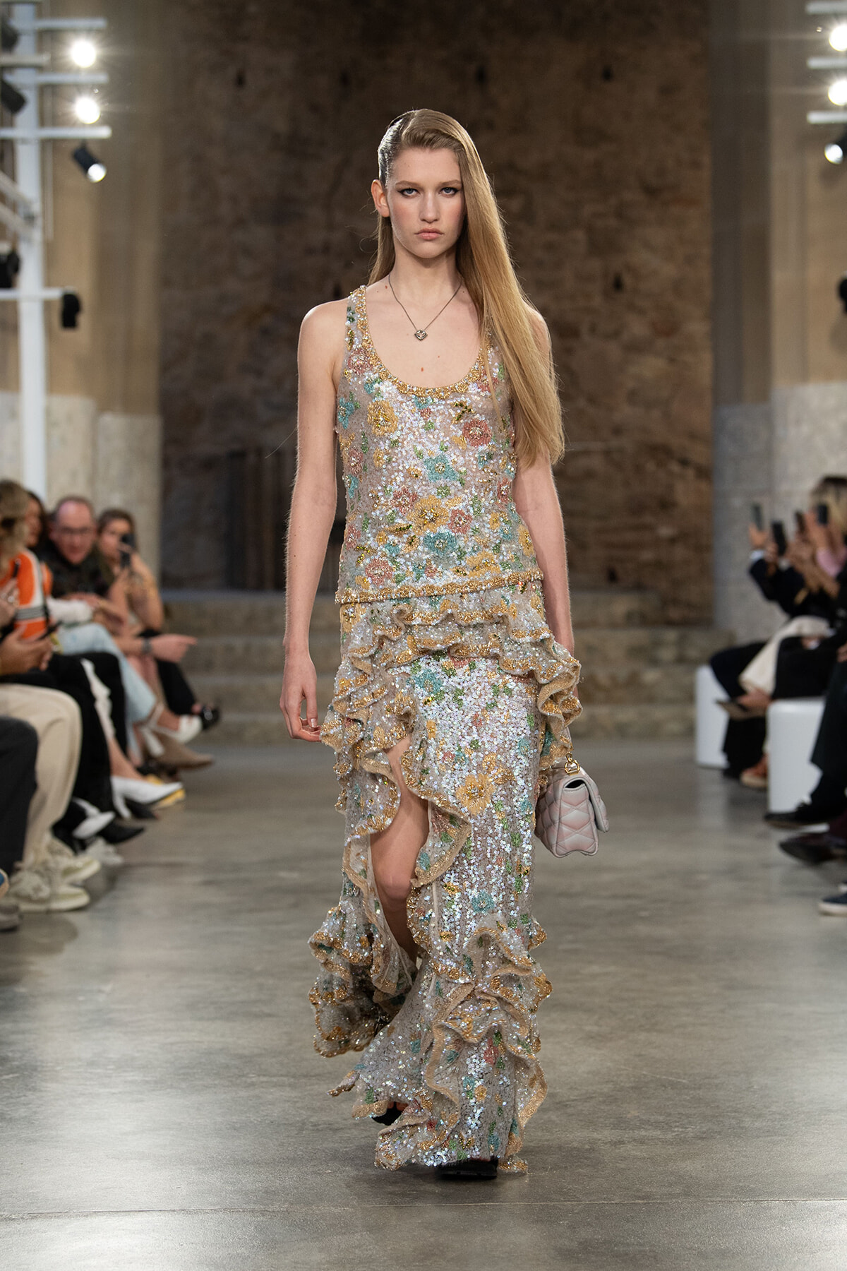 Model on a runway wearing a long, glittering floral sequin gown with cascading ruffles and a pale pink quilted purse.