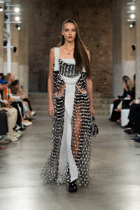 Model walks the runway in a black-and-silver sequined gown with sheer dotted panels and a white belt-like sash, at a fashion show.