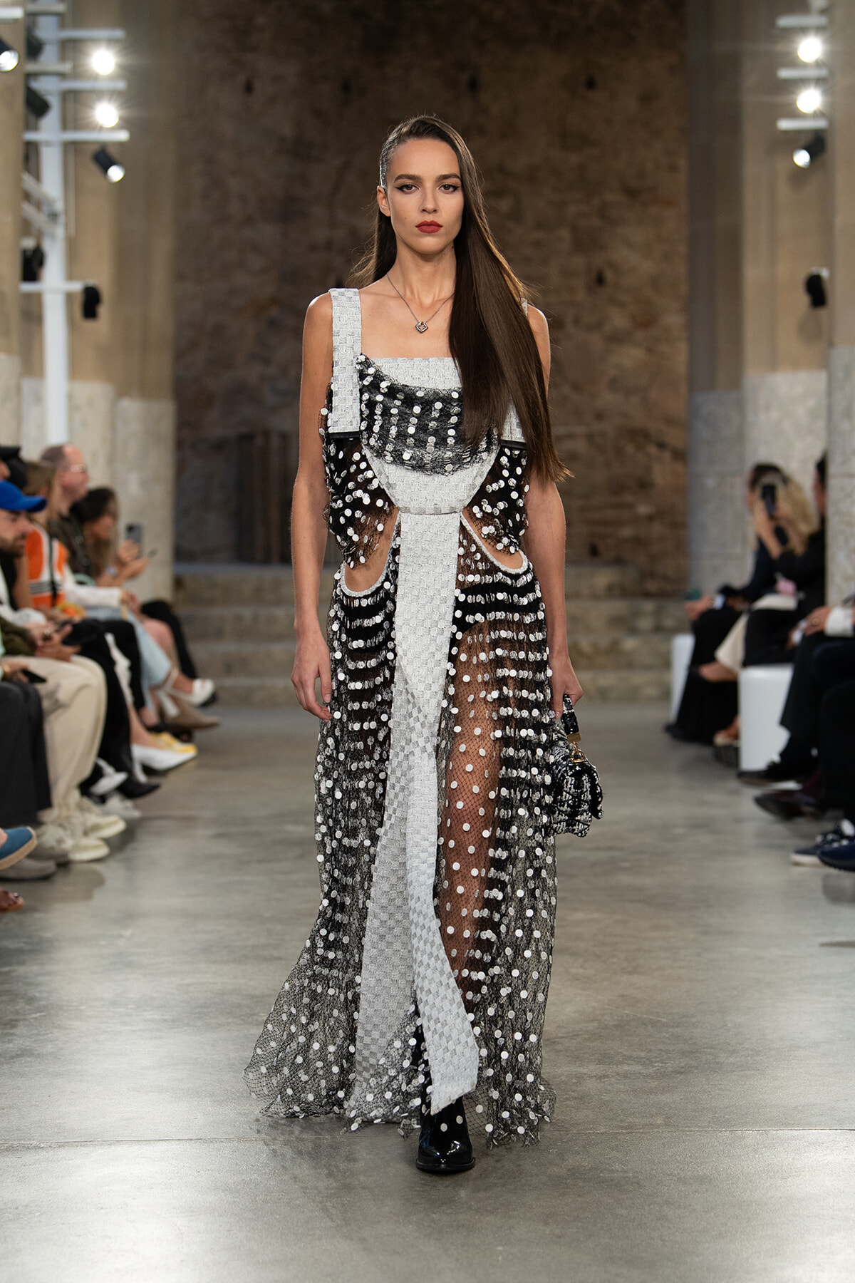 Model walks the runway in a black-and-silver sequined gown with sheer dotted panels and a white belt-like sash, at a fashion show.