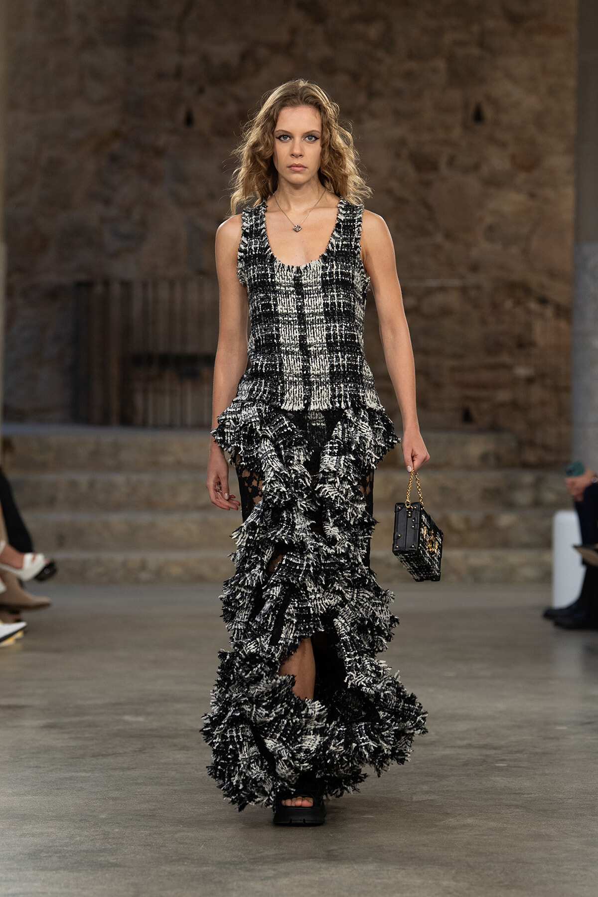 Model walking a runway in a black-and-white tweed gown with multiple layered ruffles and a high-low hem, carrying a small black handbag with a chain strap.