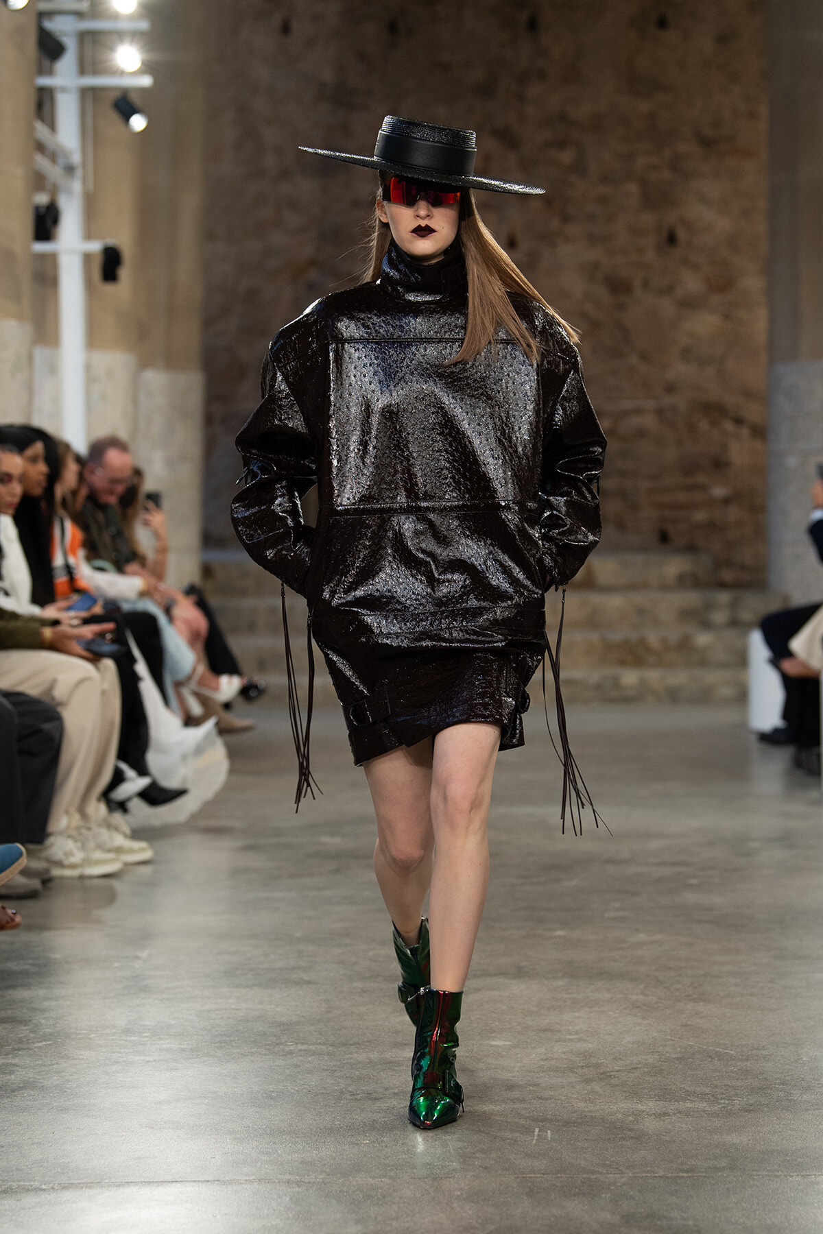Model struts the runway in a glossy black oversized top and skirt, wide-brim black hat, red visor sunglasses, and shiny green ankle boots on a fashion show runway.
