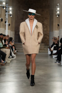 Runway model in a beige textured coat with oversized white lapels, white hat, teal sunglasses, and black ankle boots.