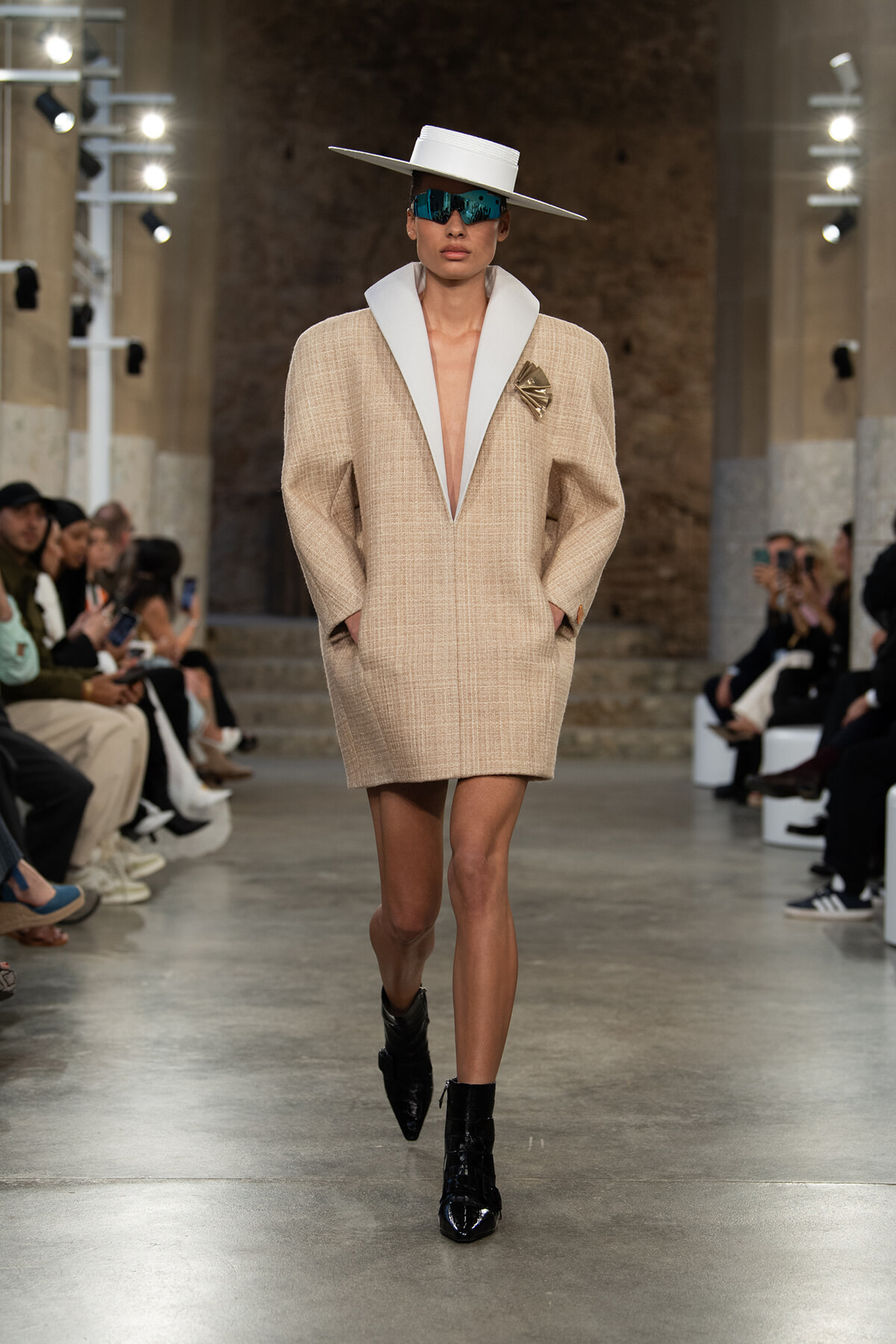 Runway model in a beige textured coat with oversized white lapels, white hat, teal sunglasses, and black ankle boots.