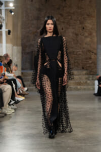 Model walking a runway in a long black sheer polka-dot lace dress with cape-like sleeves in an industrial venue.