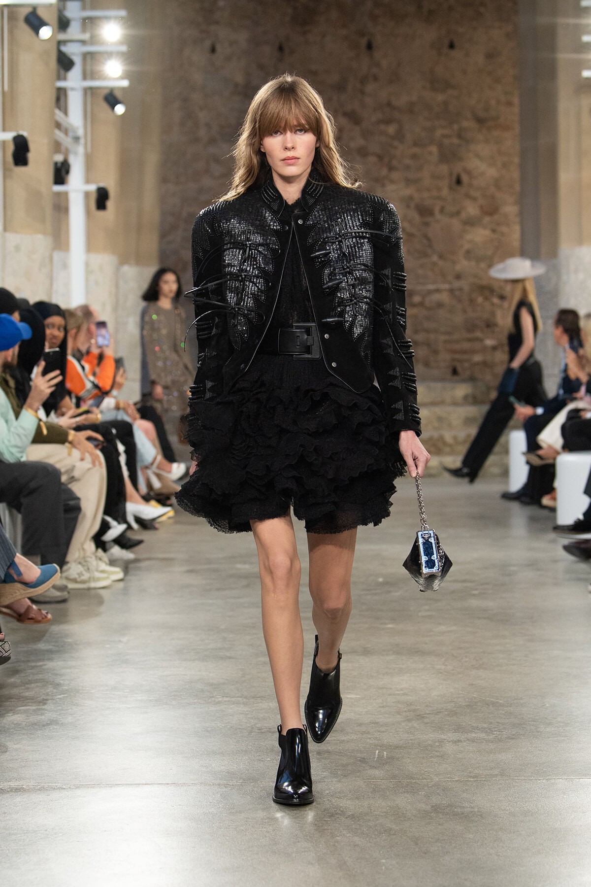 Runway model in a black textured jacket with a voluminous layered ruffled skirt, carrying a small geometric handbag.