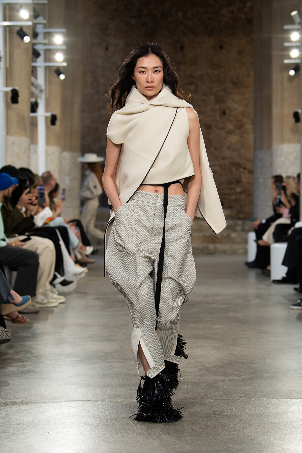 Model walking a fashion runway in a cream asymmetrical cape and loose patterned pants with a black belt, paired with fringed black boots.