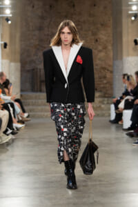 Runway model wearing a black blazer with white lapels, red pocket square, mosaic-print trousers, black boots, and a black chain-strap handbag.