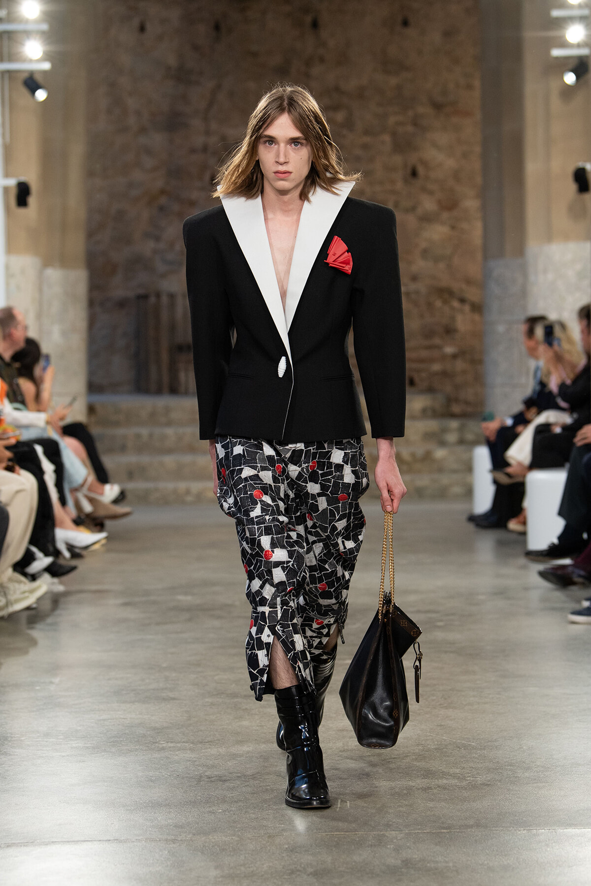 Runway model wearing a black blazer with white lapels, red pocket square, mosaic-print trousers, black boots, and a black chain-strap handbag.