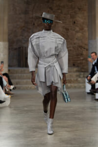 Runway model in a structured light-gray dress with a belt, oversized sleeves, and a high collar, paired with white ankle boots and a teal visor hat, carrying a small blue handbag.
