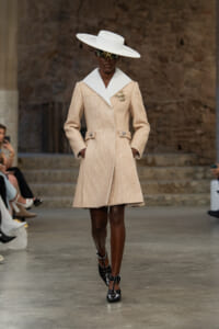 Model walks the runway in a structured beige coat with large white lapels, a gold brooch, black heeled shoes, and a wide white hat.
