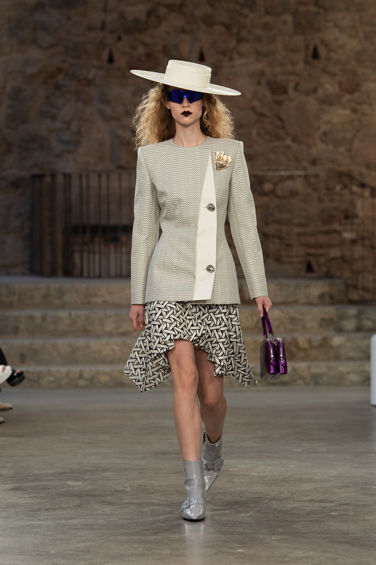 Runway model in a structured gray check blazer with a white diagonal panel, geometric skirt, white wide-brim hat, blue sunglasses, gray boots, and a purple handbag.