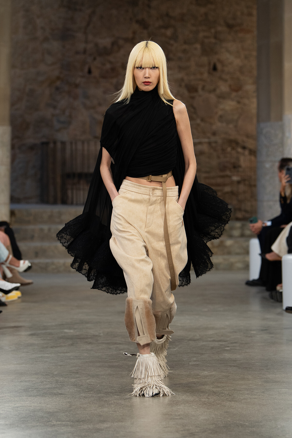 Runway model walks the catwalk in a black draped top, beige cargo pants, and fringed boots with platinum blonde hair — fashion show.