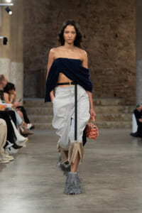 Model walks a fashion runway wearing a navy draped off-the-shoulder top, light gray wide pants, beige fringe boots, and a pink quilted handbag.