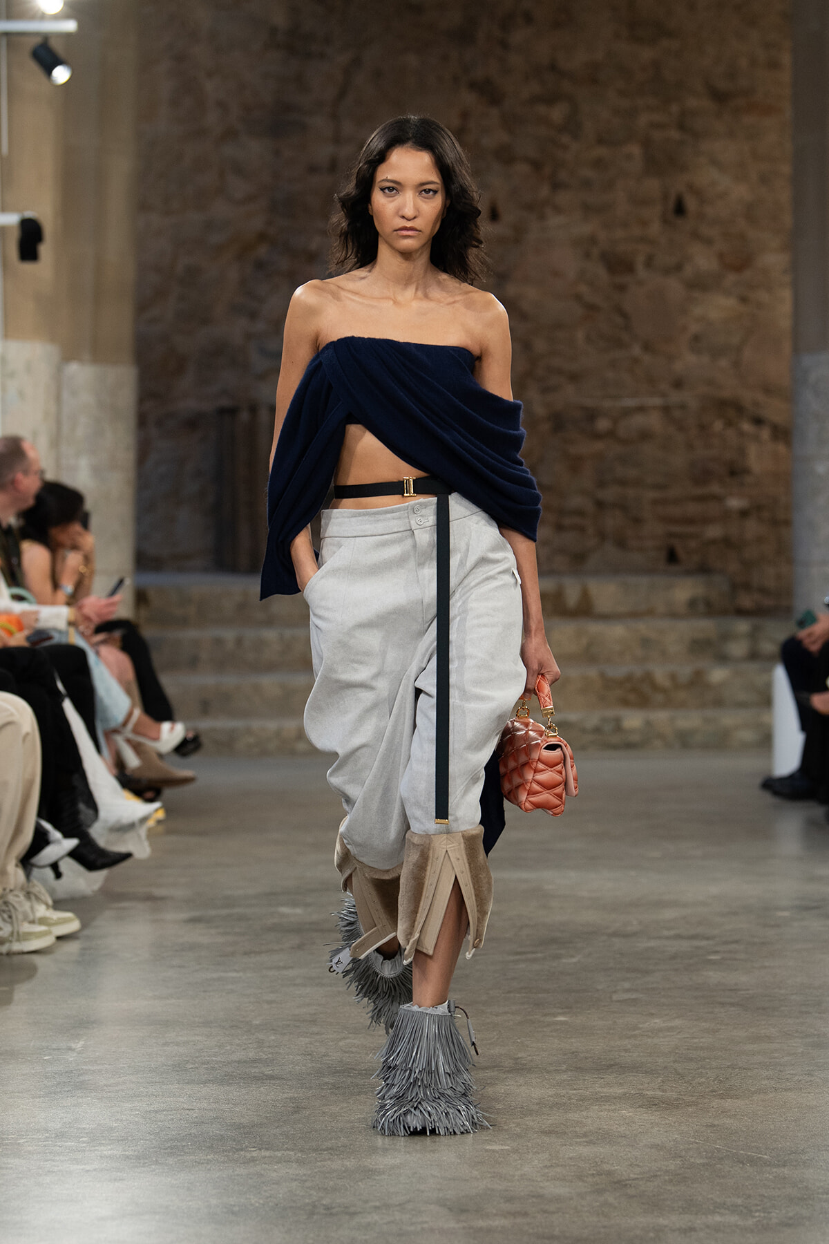 Model walks a fashion runway wearing a navy draped off-the-shoulder top, light gray wide pants, beige fringe boots, and a pink quilted handbag.