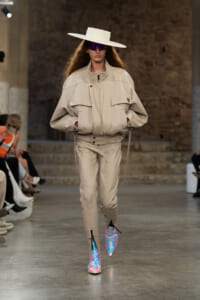 Runway model in a beige oversized jacket and pants, wide-brim white hat, purple reflective sunglasses, and iridescent boots.