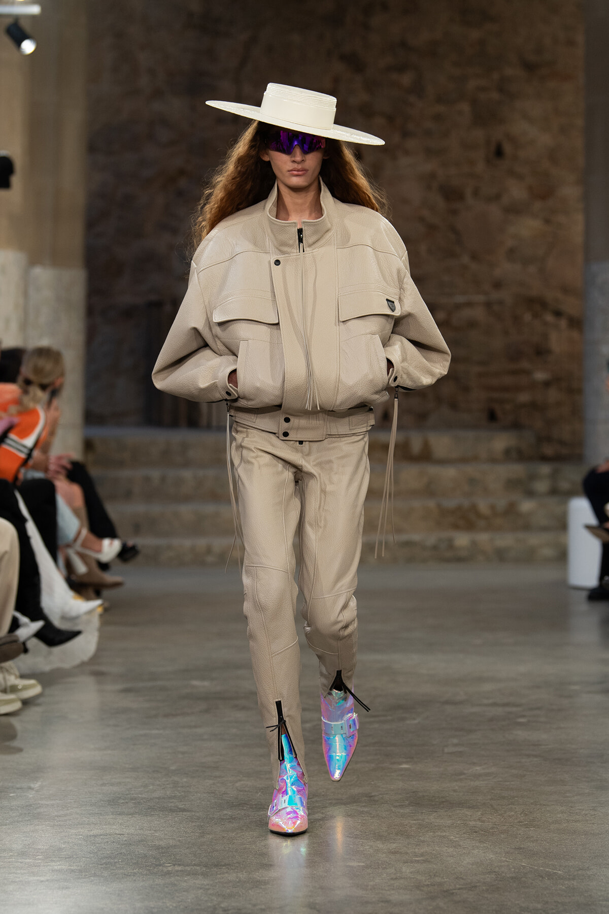 Runway model in a beige oversized jacket and pants, wide-brim white hat, purple reflective sunglasses, and iridescent boots.