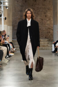 Runway model strides down the catwalk in a black long coat over a white shirt and beige pants, carrying a brown chain-strap tote.