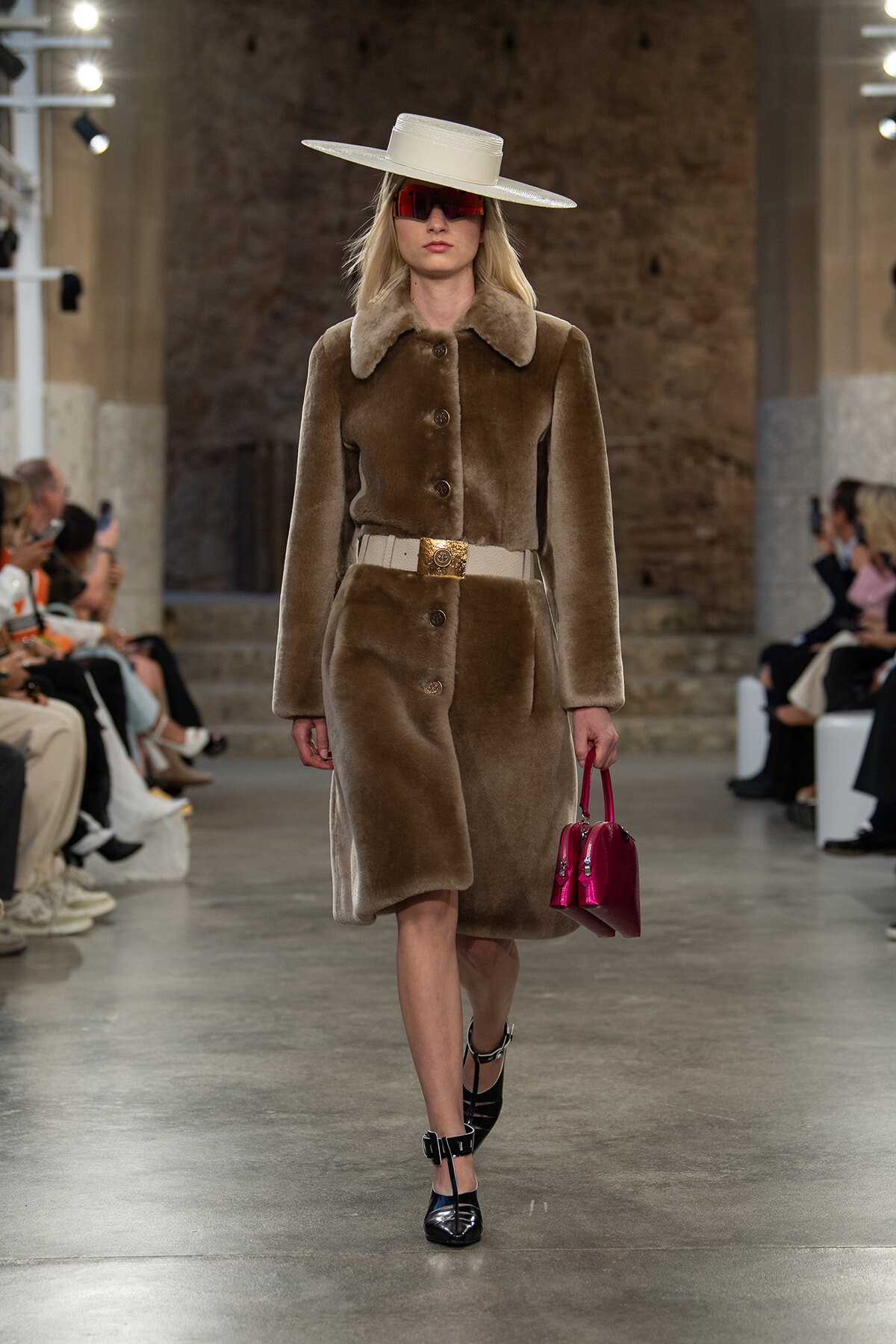 Model walking runway in a brown fur coat with a wide-brim hat, red visor sunglasses, gold belt, and bright pink handbag.