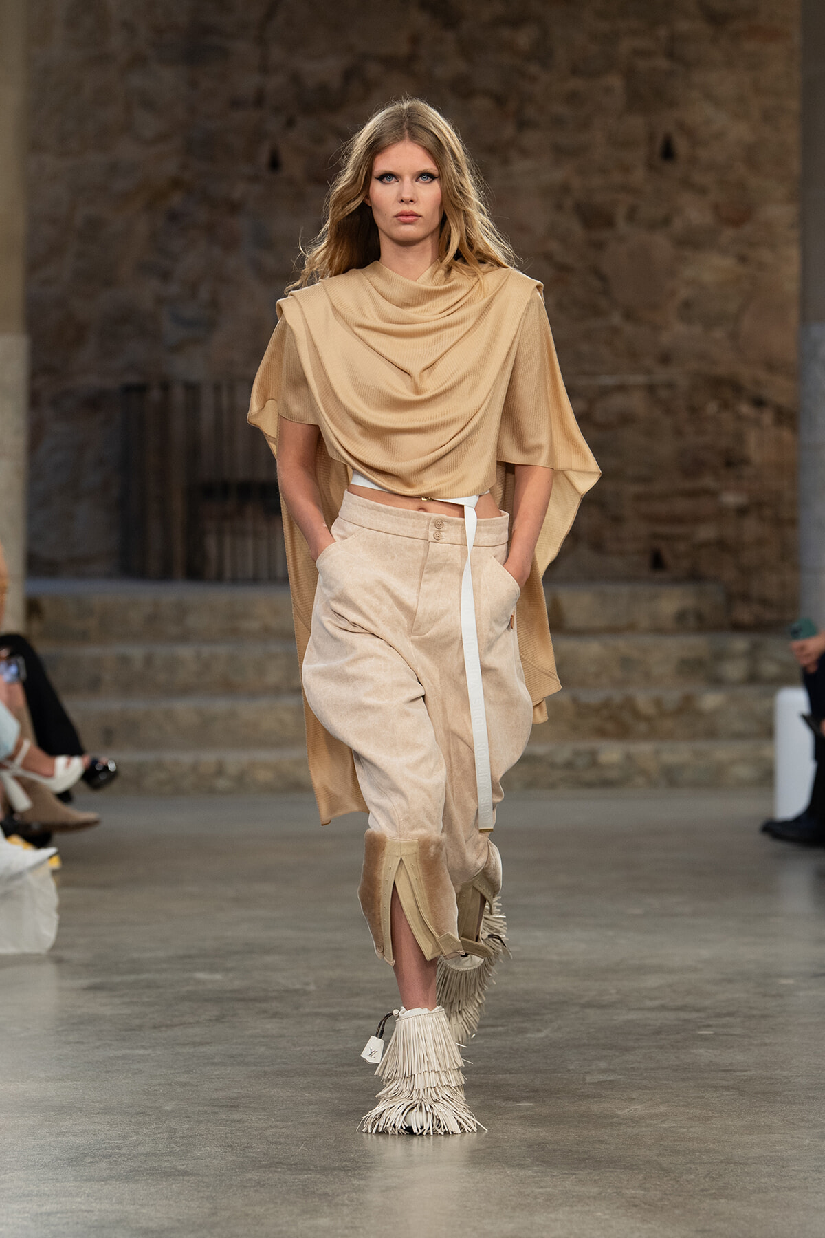 Model walks a fashion runway wearing a beige draped top, high-waisted beige pants, and fringed boots, with a stone backdrop behind.