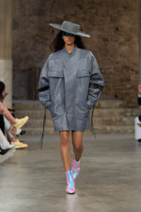 Model walking runway in oversized gray textured coat with large front pockets, iridescent pink boots, wide-brimmed hat and futuristic sunglasses.