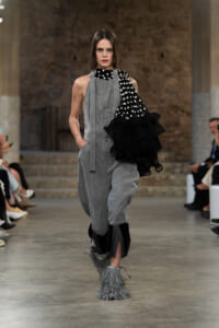 Model walks the runway in a gray utility jumpsuit with a black polka-dot one-shoulder top and a voluminous black ruffle sleeve; gray fringe boots enhance the look.
