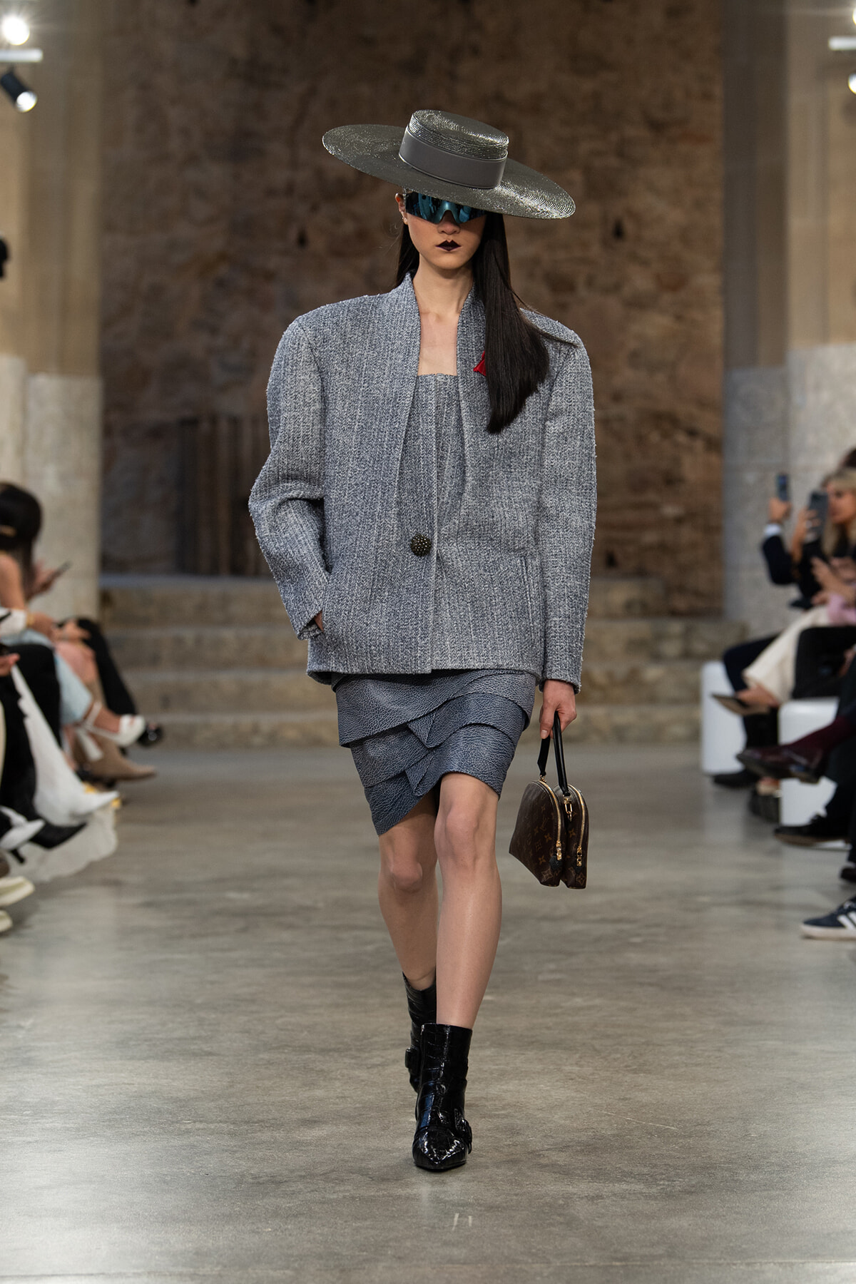Model walking a runway in a grey textured blazer and layered matching skirt, wide-brimmed hat, oversized sunglasses, and black ankle boots, carrying a brown handbag