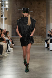 Runway model wearing a black sculptural dress with a high collar, wide belt, oversized pockets, a wide-brim black hat, green mirrored sunglasses, and iridescent green–red boots.