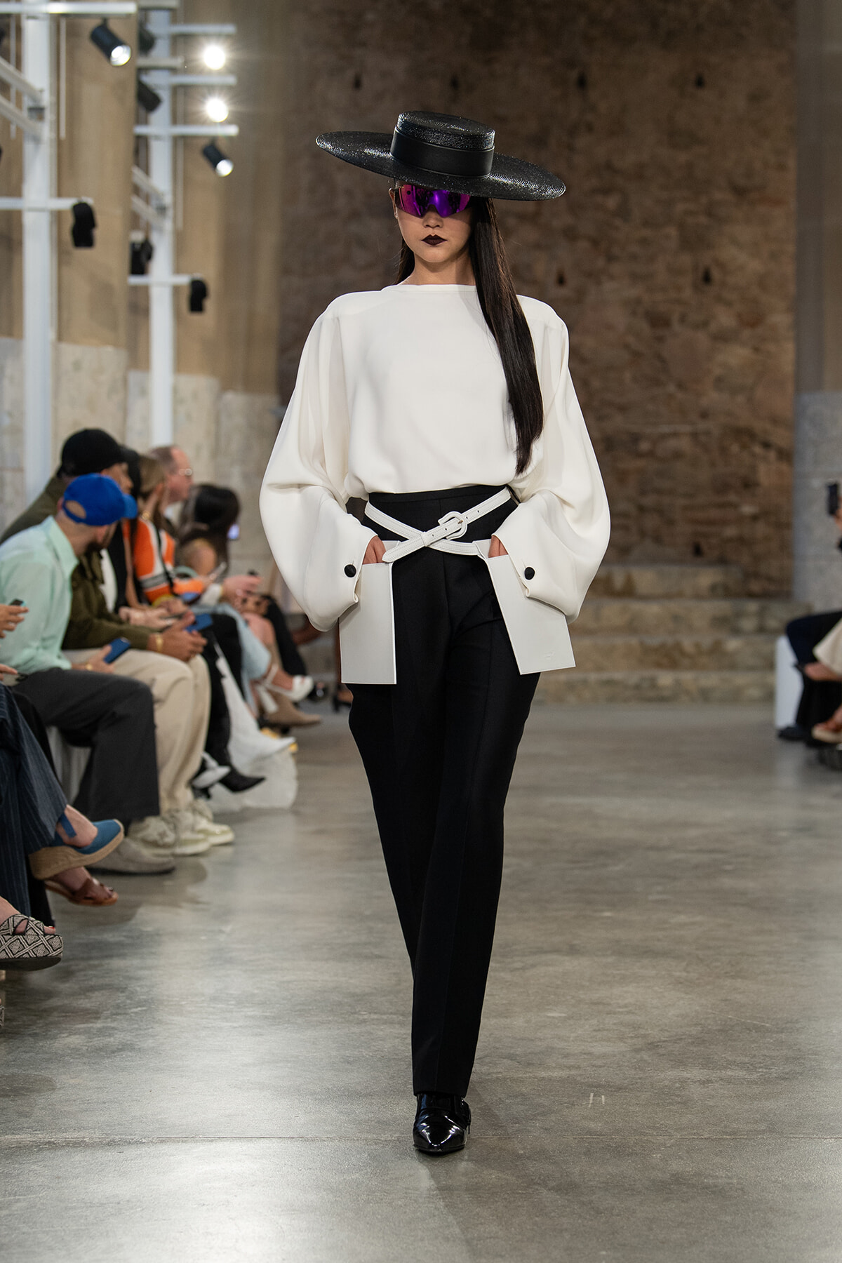 Model walking a runway in a white oversized blouse with black trousers and a white belt, wearing a wide black hat and purple sunglasses.