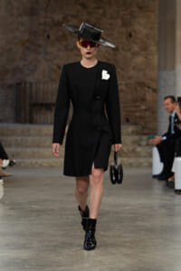 Model walks a runway in a knee-length black coat with a white corsage, wide-brim black hat, red-tinted sunglasses, and glossy black ankle boots, while carrying a small black handbag.