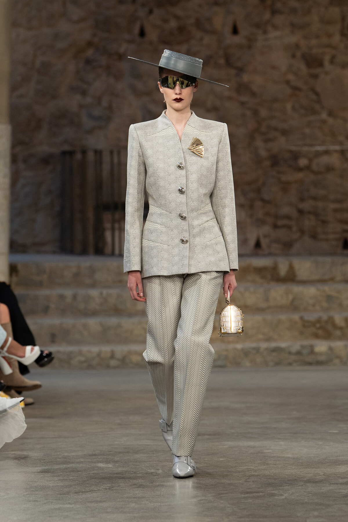 Model wearing a light gray tailored suit with structured shoulders, matching pants, metallic hat, oversized sunglasses, and a gold brooch; holding a small gold handbag.