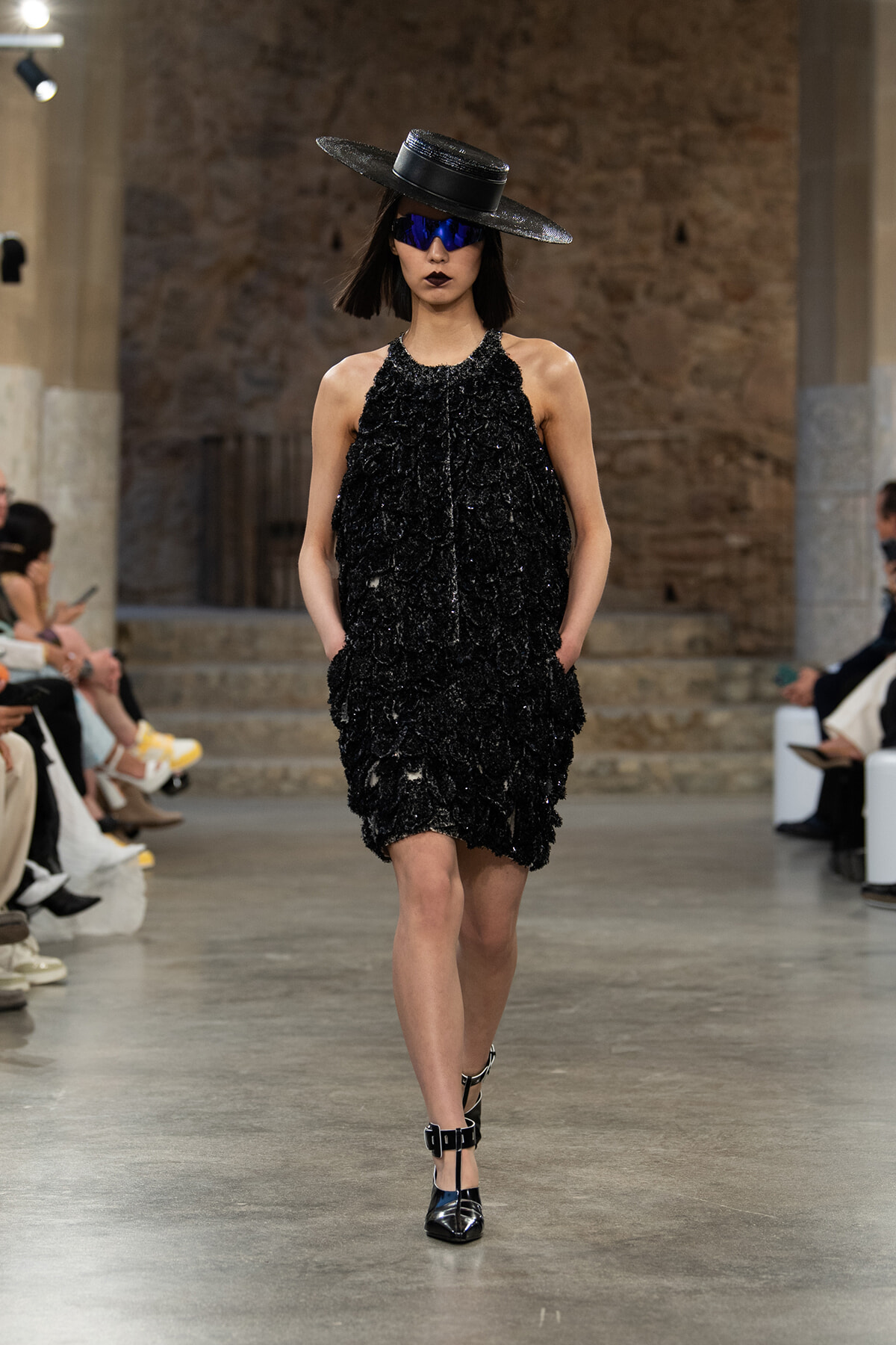 Model walks the runway in a black textured mini dress with a wide-brim hat, blue sunglasses, and strappy heels.