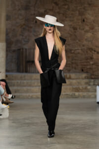 Model on a runway wearing a black plunging jumpsuit with oversized pockets, a white wide-brim hat, and mirrored blue sunglasses.