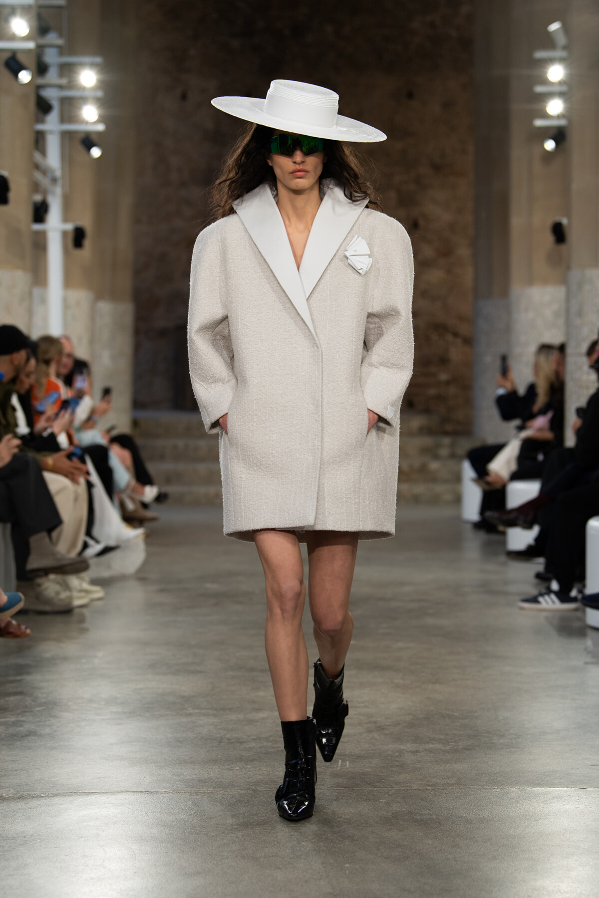 Model walking a runway in a textured oversized white coat with wide lapels, a large white hat, green sunglasses, and black ankle boots.
