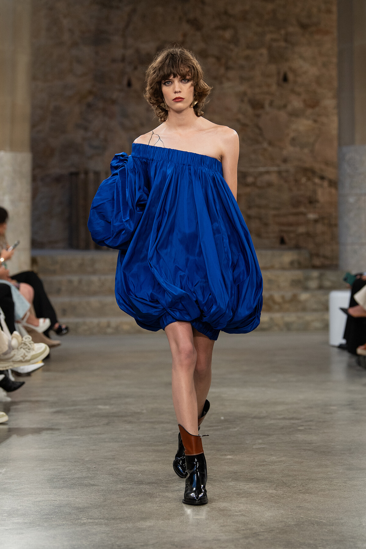 Model walking a fashion runway in a bright royal blue off-the-shoulder baby-doll dress with voluminous gathered fabric, paired with black ankle boots and brown-heeled accents.