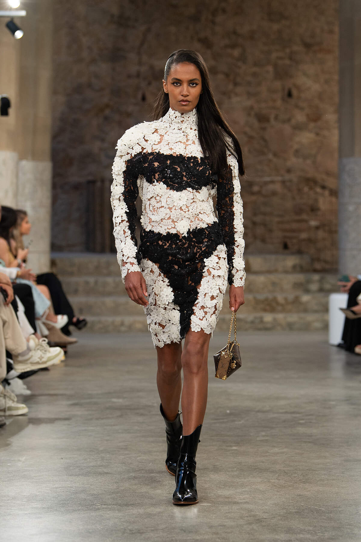 Model walking a runway in a white and black lace dress with a high neck, wearing black ankle boots, and carrying a small brown handbag with a gold chain.