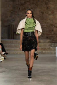 Runway model in a lime-green draped top, cream cape, black lace mini with a wide belt, and ankle boots, carrying a small handbag.