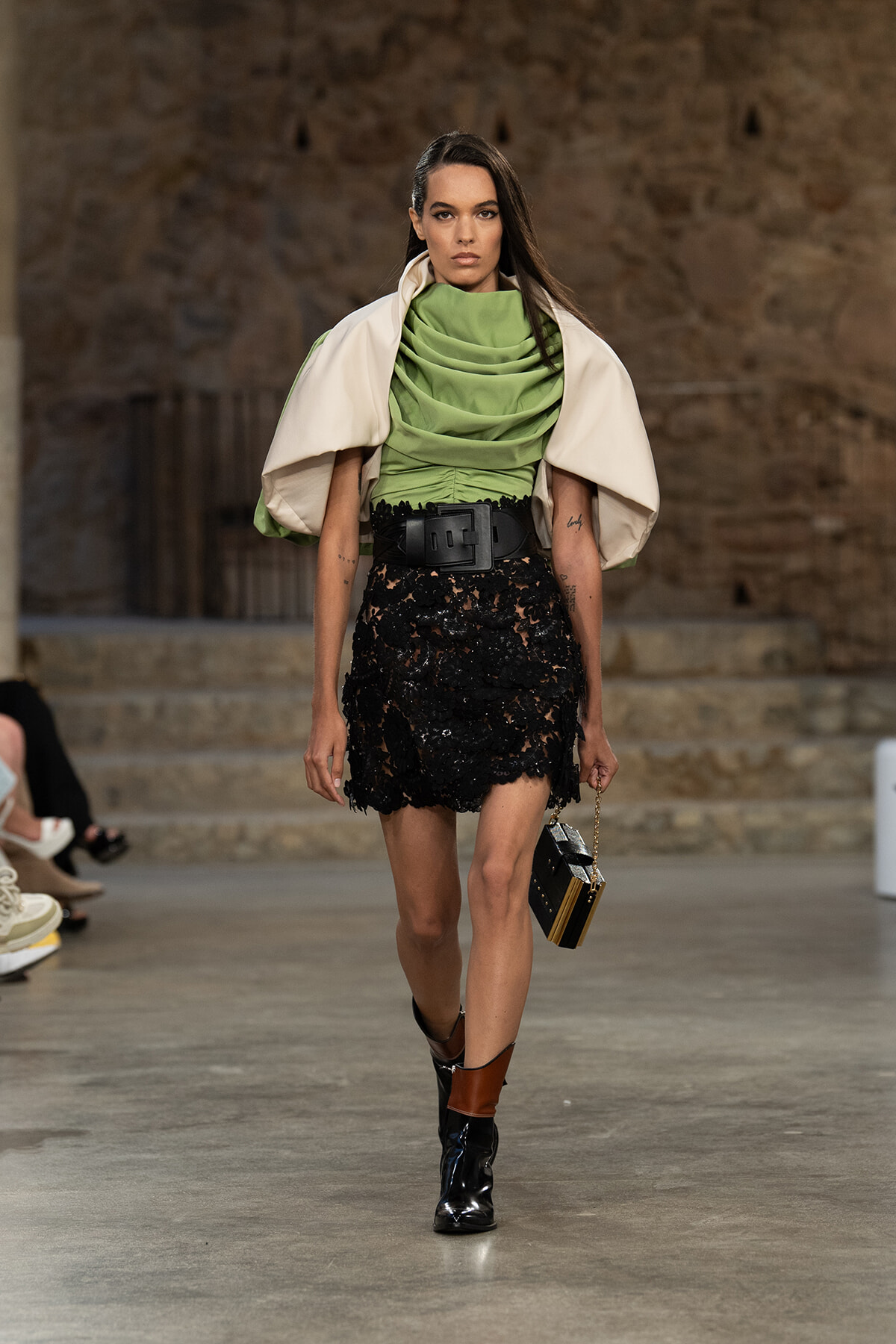 Runway model in a lime-green draped top, cream cape, black lace mini with a wide belt, and ankle boots, carrying a small handbag.