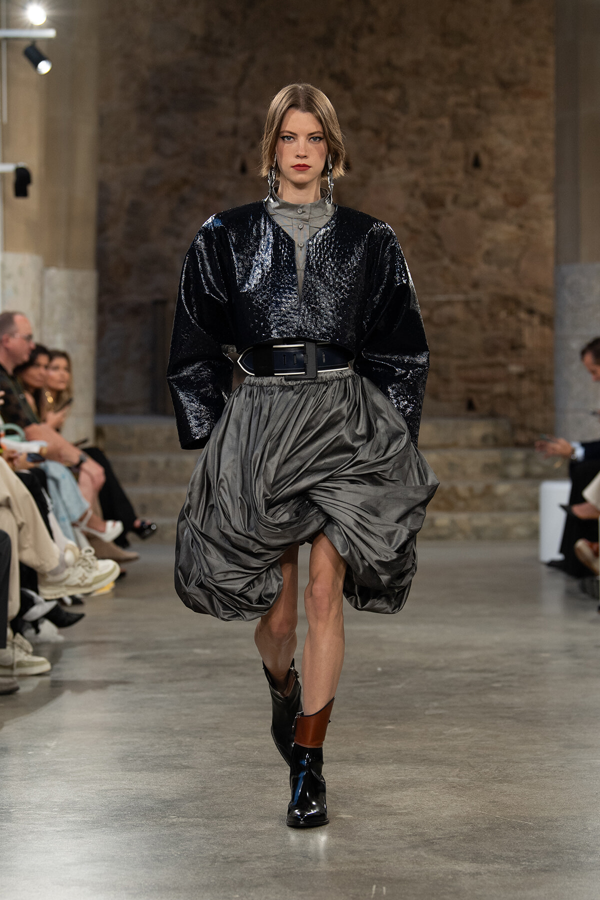 Model walks the runway in a glossy black oversized top with voluminous gray draped skirt, cinched by a wide belt and black-brown ankle boots.