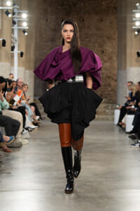 Model walks a fashion show wearing a dramatic purple draped top with a voluminous black skirt, white belt, and color-block knee-high boots.